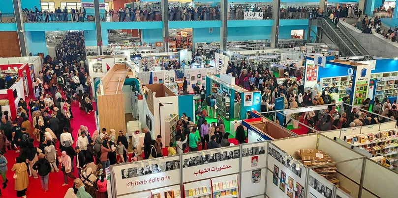 Aerial view of a crowded book fair, with numerous exhibitors and attendees moving through brightly colored booths and displays.